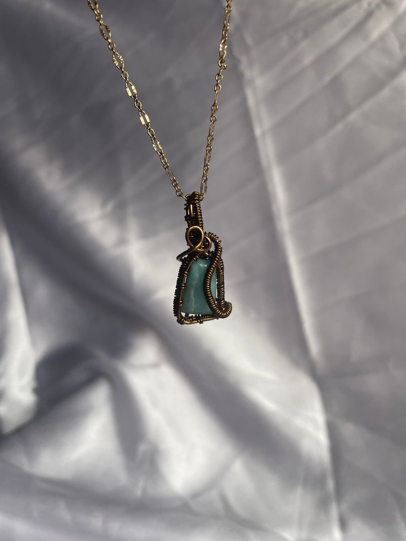 K&ouml;nnte beinhalten: Eine handgefertigte Anh&auml;ngerhalskette mit einem hellblauen Edelstein, der in bronzefarbenen Draht gewickelt ist. Der Anh&auml;nger h&auml;ngt an einer zarten goldfarbenen Kette. Die Halskette wird vor einem wei&szlig;en, strukturierten Hintergrund pr&auml;sentiert.