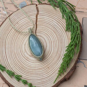 Puede incluir: Un collar con colgante hecho a mano con una gema grande, ovalada y azul pálido. La piedra está engastada en un diseño de macramé beige tejido y cuelga de un cordón a juego. El collar descansa sobre una rodaja de madera, con un mapa y ramitas verdes.