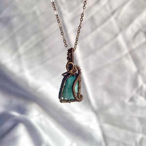 K&ouml;nnte beinhalten: Eine handgefertigte Anh&auml;ngerkette mit einem hellblauen Edelstein, der in Kupferdraht gewickelt ist. Der Anh&auml;nger h&auml;ngt an einer zarten Kupferkette. Der Edelstein hat eine weiche, opake blaugr&uuml;ne Farbe. Die Drahtarbeit weist komplizierte Details auf.