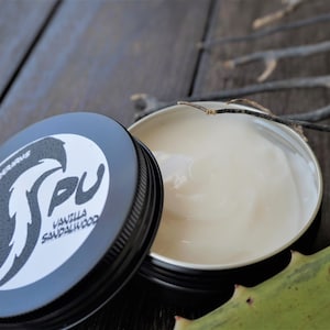 May include: Open black tin of vanilla sandalwood cream with the lid beside it. The lid has a white label with black text and a stylized bird graphic. The cream is light beige. The background is a dark wood surface.