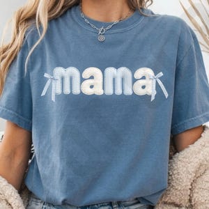 以下が含まれることがあります： 白と青のギンガムチェック柄の文字で「mama」と書かれた青い半袖Tシャツ。最初の文字と最後の文字の両端には、白と青のギンガムチェック柄の小さなリボンがあしらわれています。クルーネックのTシャツです。