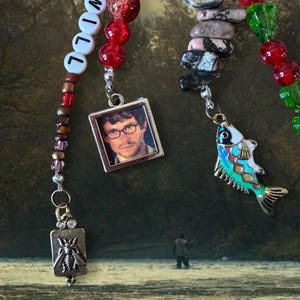 Puede incluir: Un marcapáginas de cuentas con un dije cuadrado que muestra el retrato de un hombre con gafas, un dije rectangular con una abeja y un dije con forma de pez. El cordón incluye cuentas rojas, cuentas blancas con letras que forman WILL y piedras grises.