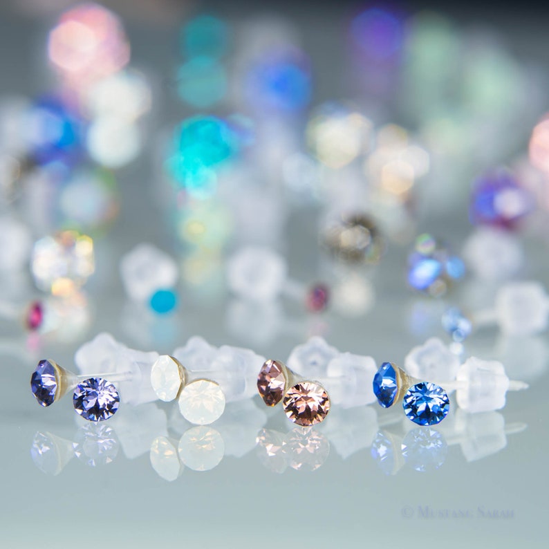 May include: Close-up of several pairs of stud earrings with various colored gemstones, including purple, white, and pink. The earrings are set on clear plastic earring backs, with a blurred background of additional earrings.