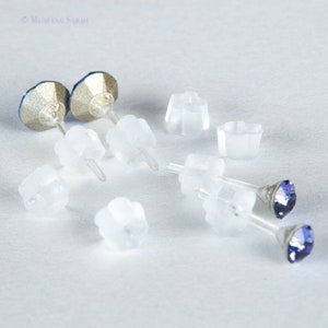 May include: A pair of blue crystal stud earrings with clear plastic earring backs. The earrings have a silver-colored base and a faceted blue crystal. Several clear plastic earring backs are scattered around the earrings.