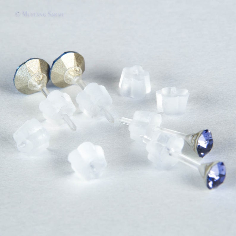 May include: A pair of blue crystal stud earrings with clear plastic earring backs. The earrings have a silver-colored base and a faceted blue crystal. Several clear plastic earring backs are scattered around the earrings.