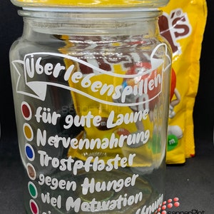 Puede incluir: Un tarro de vidrio transparente con una etiqueta blanca que dice "Überlebenspillen" y enumera las razones para comer los dulces que contiene, como "für gute Laune" (para el buen humor) y "Nervennahrung" (alimento para los nervios).