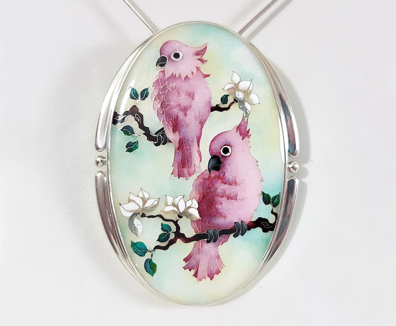 Op de afbeelding: Ovale hanger met een zilveren frame met twee roze kaketoes die op een tak zitten met witte bloemen en groene bladeren. De vogels hebben zwarte ogen en snavels, tegen een lichtblauwe en groene achtergrond.