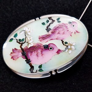 Op de afbeelding: Ovale hanger met een zilveren frame. De hanger toont twee roze vogels die op een tak zitten met witte bloemen en groene bladeren tegen een lichtgroene achtergrond. De vogels hebben zwarte ogen en zijn gedetailleerd met roze en paarse tinten.