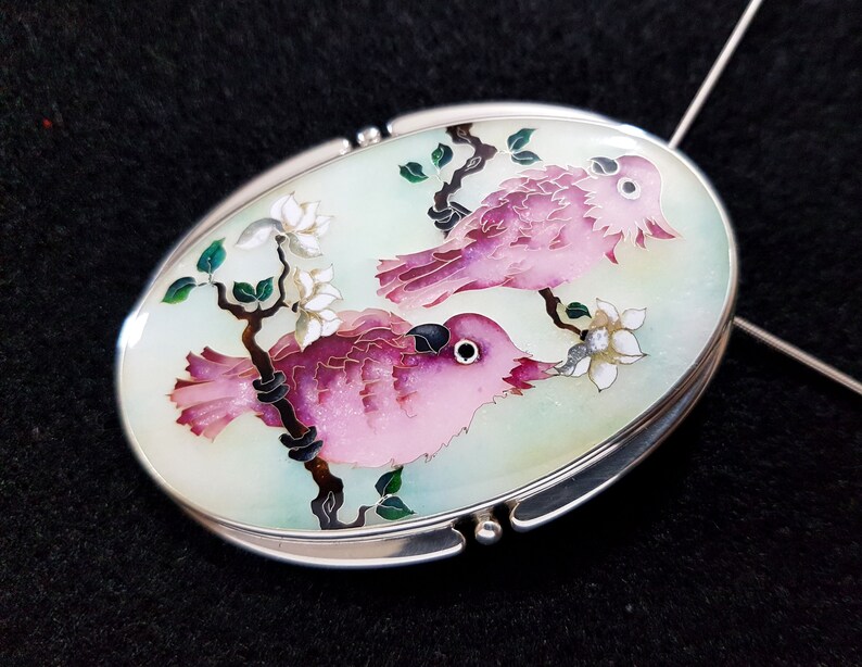 Op de afbeelding: Ovale hanger met een zilveren frame. De hanger toont twee roze vogels die op een tak zitten met witte bloemen en groene bladeren tegen een lichtgroene achtergrond. De vogels hebben zwarte ogen en zijn gedetailleerd met roze en paarse tinten.
