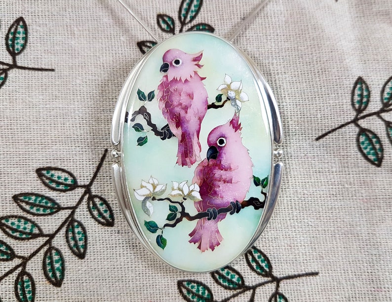 Op de afbeelding: Ovale hanger met een zilveren frame met een aquarel van twee roze kaketoes op een tak met witte bloemen. De achtergrond is lichtgroen.