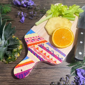 Pu&ograve; includere: Un tagliere di legno colorato con un motivo geometrico. Il tagliere ha un manico ed &egrave; decorato con colori vivaci rosa, arancio, giallo, blu e verde. Ci sono un ananas, un'arancia e sedano sul tagliere.