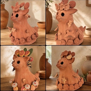 May include: A terracotta cat figurine adorned with various sculpted clay elements including flowers, butterflies, and small skulls. The cat has large circular eye cutouts, thin wire whiskers, and a textured surface decorated with floral motifs.