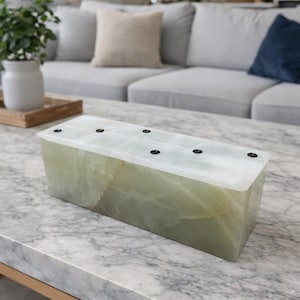 May include: A rectangular, light green onyx vase with a white top featuring six black circular openings. The vase is set on a marble-topped coffee table. The overall aesthetic is modern and minimalist.