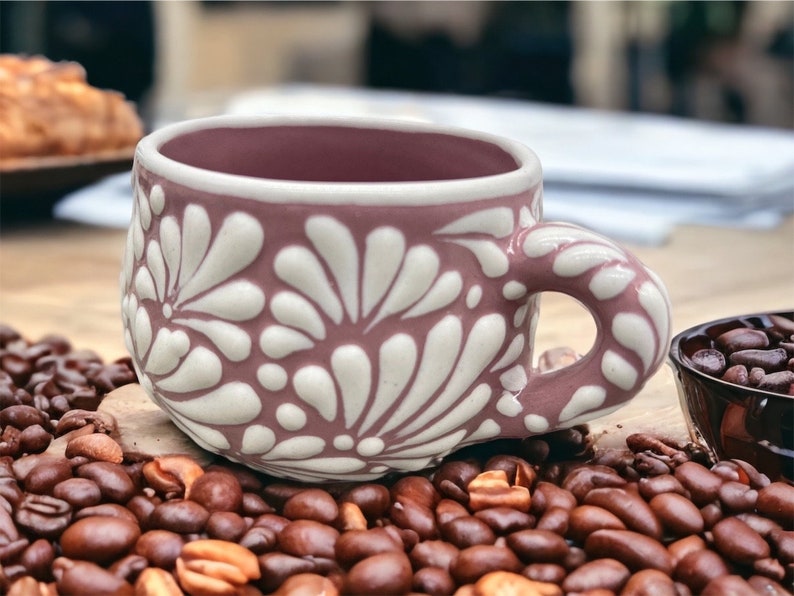 May include: A pink ceramic coffee cup with a white floral design. The cup is sitting on a bed of coffee beans.