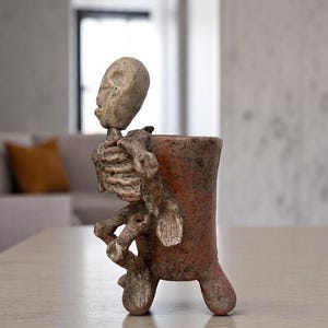 Op de afbeelding: Een handgemaakte keramische plantenbak met een skeletfiguur. De roodbruine plantenbak heeft een textuur en het skelet is lichtbeige. Het skelet lijkt de plantenbak te omarmen. De plantenbak heeft drie poten.