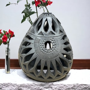 May include: A gray ceramic vase with intricate cutouts and a circular design in the center. The vase is sitting on a white surface with a small vase of red flowers in the background.
