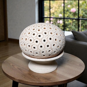 Op de afbeelding: Een crèmekleurig, bolvormig decoratief object met een cirkelvormig patroon van gaten, rustend op een witte sokkel. Het object staat op een ronde, houten tafel. De achtergrond omvat een raam en een glimp van een bank.