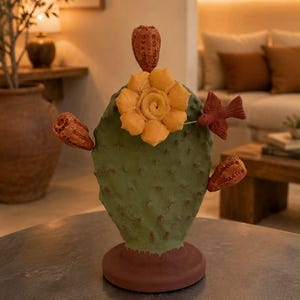 May include: A decorative ceramic cactus sculpture featuring a textured green pad, a large yellow flower at the center, three reddish-brown prickly pear fruits, and a small reddish-brown bird perched on the side, all set on a circular base.