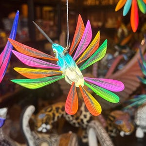 May include: A colorful, handcrafted hummingbird ornament with a long beak and outstretched wings. The wings are painted in vibrant shades of pink, orange, yellow, and green. The bird's body is a mix of blue and yellow. The ornament hangs from a thin string.