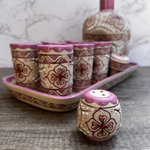Puede incluir: Un juego de licor de cer&aacute;mica decorativo con una botella, vasos de chupito y un salero. El juego tiene una base beige con detalles en rosa y dise&ntilde;os florales. Los vasos de chupito est&aacute;n colocados en una bandeja a juego.