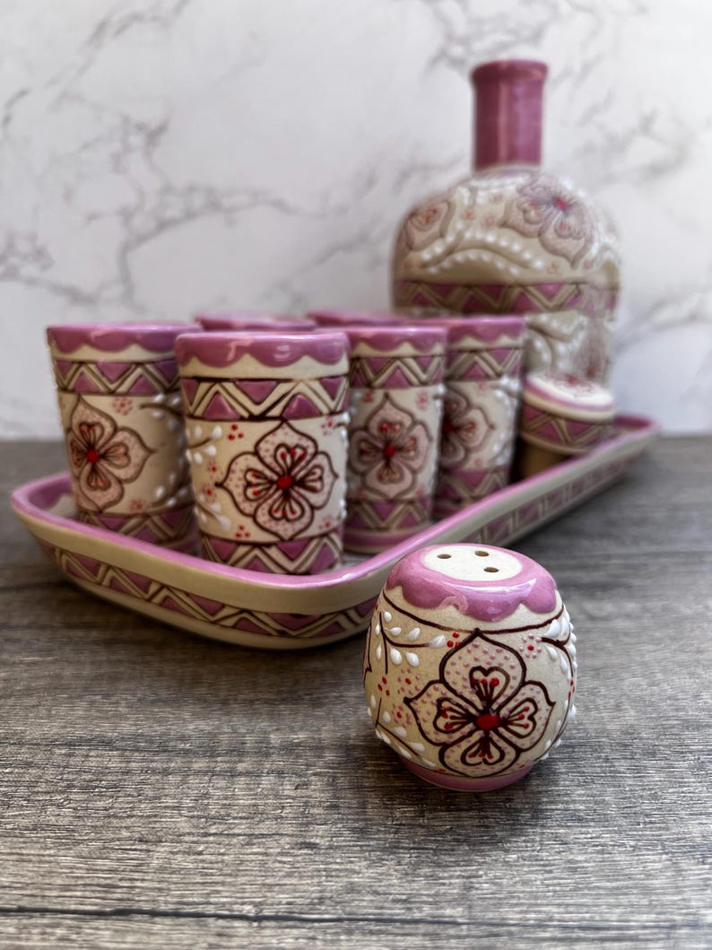 Puede incluir: Un juego de licor de cer&aacute;mica decorativo con una botella, vasos de chupito y un salero. El juego tiene una base beige con detalles en rosa y dise&ntilde;os florales. Los vasos de chupito est&aacute;n colocados en una bandeja a juego.