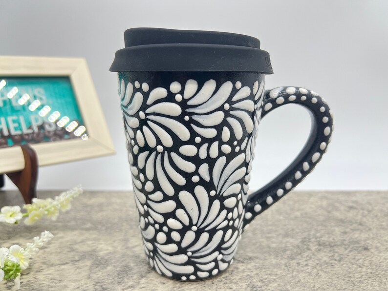 Puede incluir: Taza de viaje de cer&aacute;mica negra y blanca con un patr&oacute;n floral y una tapa de silicona negra. La taza tiene un asa con puntos blancos.