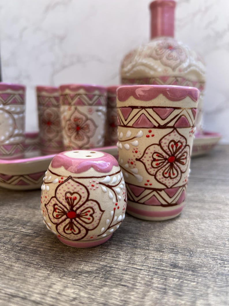Puede incluir: Un juego de vajilla de cer&aacute;mica con un salero, un vaso alto y una botella. Las piezas est&aacute;n decoradas con un estampado floral en rosa y blanco, con detalles en marr&oacute;n. El juego incluye una bandeja y varios vasos.