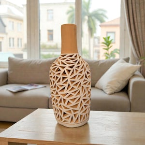 Könnte beinhalten: Eine hohe, dekorative Vase mit geometrischem Muster. Die Vase hat einen hellbraunen Hals und einen weißen Körper mit dreieckigen Aussparungen. Sie steht auf einem Holztisch in einem Wohnzimmer.