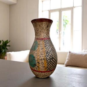 May include: A decorative vase with a speckled beige finish, featuring a red rim and base. The vase has a patterned design with teal circles, black crosshatching, and yellow accents. It is displayed on a gray surface.