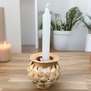 May include: A white candle sits in a ceramic candle holder with a textured, shell-like design. The holder is a light beige color.
