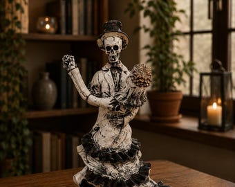 Könnte beinhalten: Eine dekorative Figur, die zwei Skelette in einer Tanzpose darstellt. Ein Skelett trägt einen Zylinder und ein Sakko, während das andere ein Kleid mit Rüschen trägt. Die Figuren haben eine Oberfläche in abgenutztem Weiß und Schwarz.