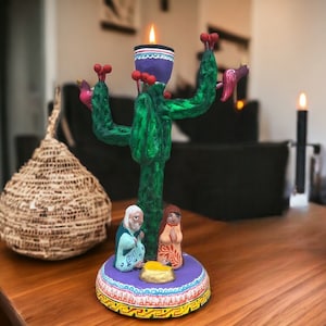 May include: A green cactus-shaped candle holder with a purple candle and three red birds. The base of the holder features a nativity scene with a brown-haired woman, a man with a white beard, and a baby in a manger.