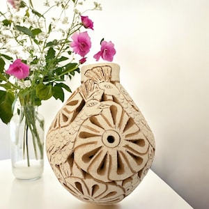 May include: A beige ceramic vase with a textured surface and intricate carvings of birds and flowers. The vase has a round body and a narrow opening at the top.