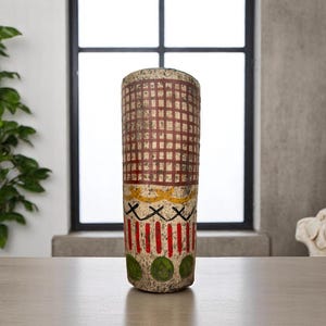 May include: A cylindrical vase with a textured surface. The upper section features a red grid pattern, followed by yellow and black designs, and vertical red stripes. Green circles adorn the base. The vase is approximately 12 inches tall.
