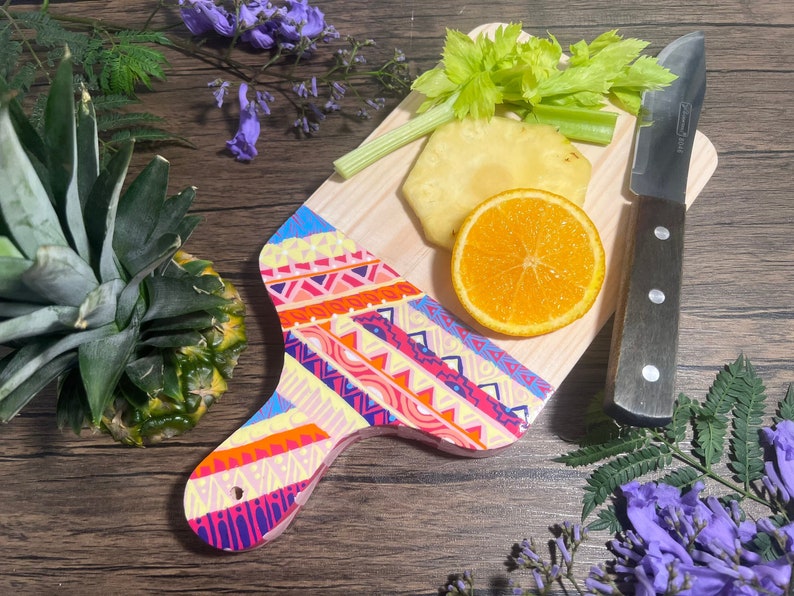Pu&ograve; includere: Un tagliere di legno colorato con un motivo geometrico. Il tagliere ha un manico ed &egrave; decorato con colori vivaci rosa, arancio, giallo, blu e verde. Ci sono un ananas, un'arancia e sedano sul tagliere.