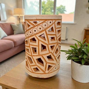 Op de afbeelding: Een cilindrische, terracotta-kleurige keramische lantaarn met geometrische uitsparingen. De lantaarn heeft een reeks driehoeken, vierkanten en cirkels die in het oppervlak zijn gesneden. De lantaarn is ongeveer 30 cm hoog en staat op een houten tafel.