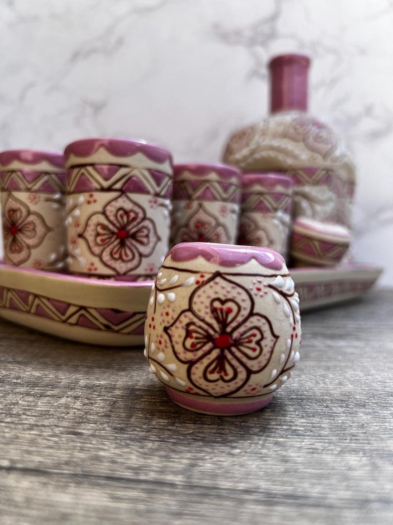 Puede incluir: Un juego de cer&aacute;mica decorativo para bebidas con una botella, vasos de chupito y una bandeja. Las piezas beige tienen detalles rosas y dise&ntilde;os florales. El conjunto se exhibe sobre una superficie de madera.