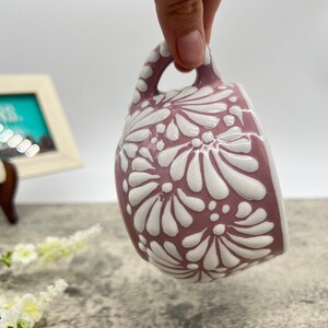 May include: A pink ceramic mug with a white floral design. The mug has a rounded shape and a handle.