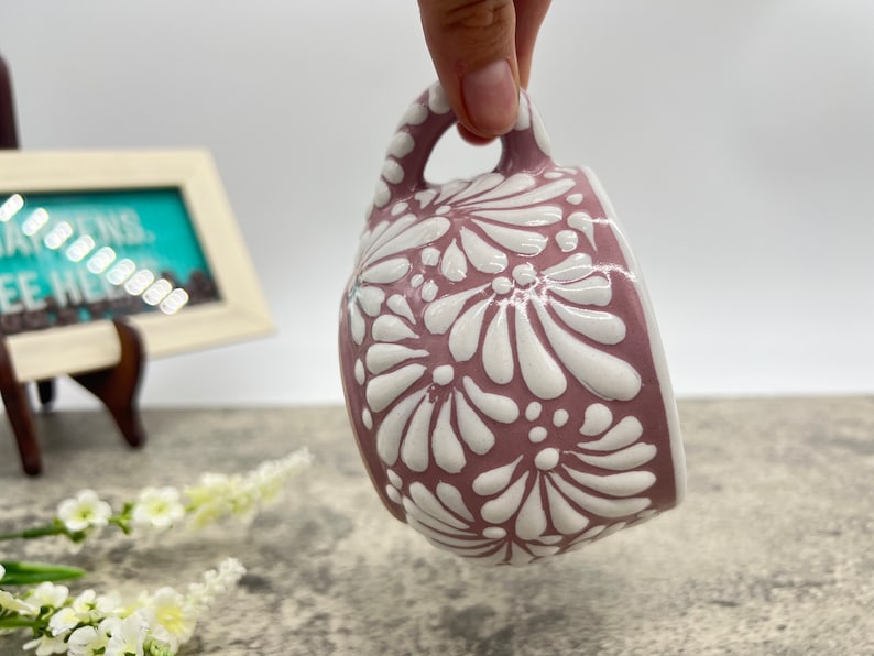 May include: A pink ceramic mug with a white floral design. The mug has a rounded shape and a handle.