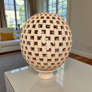 Op de afbeelding: Een bolvormig, beige decoratief object met een geweven patroon van rechthoekige openingen. De bol staat op een bijpassende beige voet, wat een uniek accent voor de woondecoratie creëert. Het ontwerp laat licht door, waardoor interessante schaduwen ontstaan.