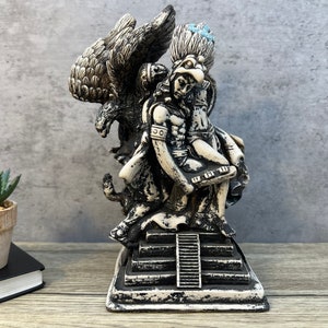 May include: A detailed, black and white statue depicting a figure with an eagle, possibly representing a deity or historical figure. The statue is mounted on a stepped base, with intricate details on the clothing and wings. The statue is approximately 8 inches tall.