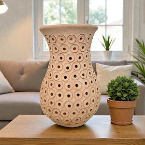 Op de afbeelding: Een beige keramische vaas met een reeks cirkelvormige uitsparingen. De vaas staat op een houten tafel. De achtergrond omvat een bank en een raam. De vaas is ongeveer 40 cm hoog.