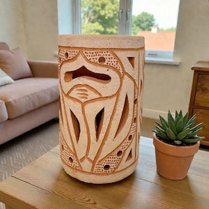 May include: A cylindrical, beige ceramic lantern with intricate carved patterns and cut-out designs. The lantern sits on a wooden table next to a small potted succulent. The background includes a sofa and a wooden cabinet.