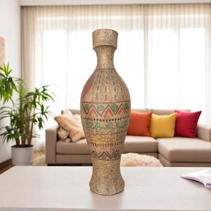 May include: A tall, beige decorative vase with a textured surface and a wide base. The vase features colorful geometric patterns in red, green, and yellow. It stands on a white table in a living room setting.