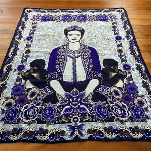 May include: A colorful throw blanket featuring a portrait of Frida Kahlo surrounded by flowers and monkeys. The blanket has a white background with a blue, purple, and gold floral pattern.