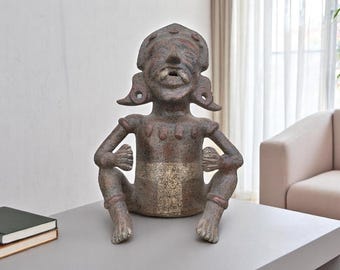 May include: A seated, weathered statue of a figure with a headdress, ear ornaments, and detailed facial features. The statue is primarily brown with reddish accents and sits on a gray surface. The sculpture's arms are bent, and its legs are crossed.