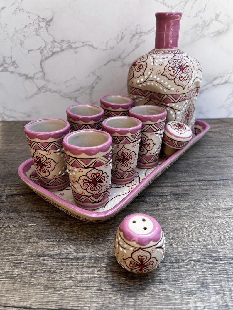 Puede incluir: Un juego de licor de cer&aacute;mica decorativo con una botella, vasos de chupito y una bandeja. El juego est&aacute; pintado con un dise&ntilde;o floral en tonos de rosa, blanco y rojo. Los vasos de chupito y la botella tienen un dise&ntilde;o a juego, y se incluye un salero.