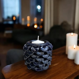May include: A dark blue ceramic candle holder with a textured, scale-like design. The holder contains a white tea light candle. The candle holder is on a wooden surface, with other lit candles in the background.