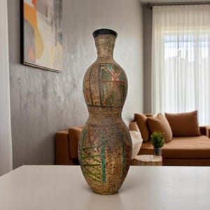 May include: A tall, decorative vase with a bulbous shape and a narrow neck. The vase is tan with green and red painted designs. The top of the vase is black. The vase is displayed on a white table in a living room.