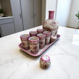 Puede incluir: Un juego de licor de cer&aacute;mica decorativo con una botella, vasos de chupito y un salero. El juego tiene un esquema de color rosa y crema con patrones florales. Los vasos de chupito y la botella est&aacute;n colocados en una bandeja a juego.